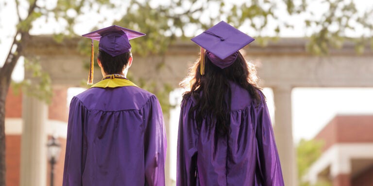 Two students wearing purple graduation gowns and caps stand with their backs to the camera, facing arches in front of them.