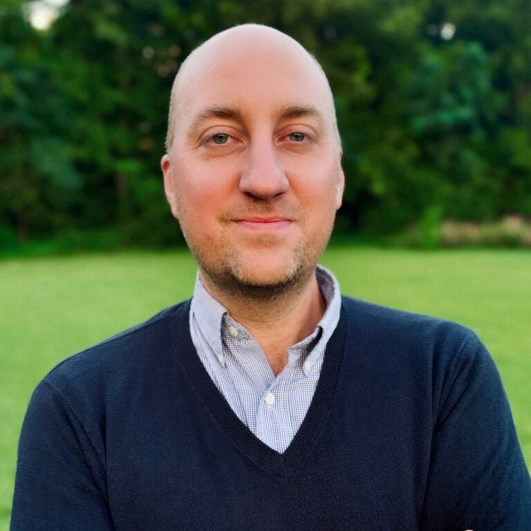 ECU alumni Dean Tuck is wearing a dark blue, v-neck sweater and button-down shirt, and is standing outside with greenery in the background.