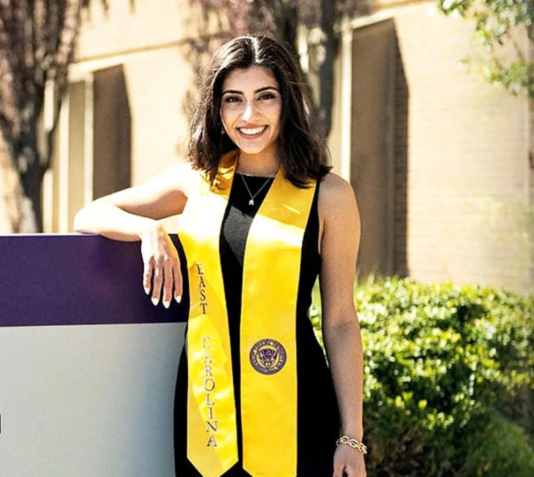 Isabella Sardina stands in front of a sign on campus in a gold graduation stole and black dress.