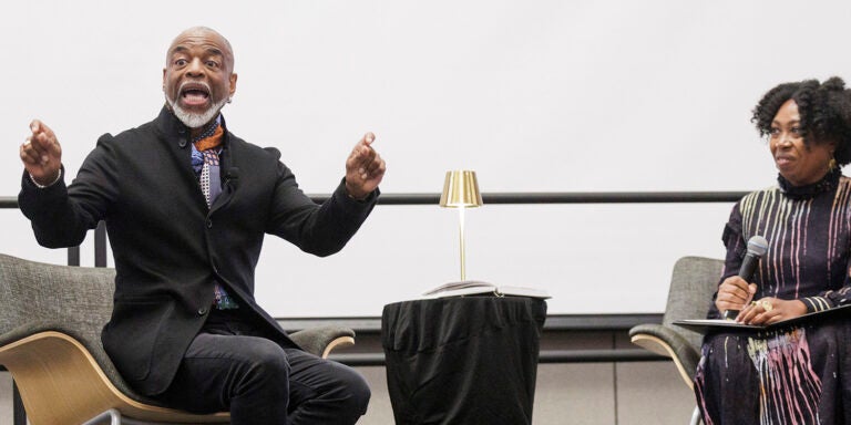 A nearly bald man with a white mustache and goatee, wearing a colorful scarf and black suit jacket, gestures with his hands while sitting to the left of a small table with a gold lamp. A woman with black, curly hair wearing a black dress with a colored streak pattern, sits to the right and holds a microphone with a folder in her lap.