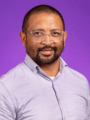 Dr. Fidisoa Rasambainarivo has short, dark hair and a mustache and beard. He is wearing clear-framed glasses and a light purple shirt in front of a purple photo backdrop.