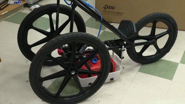 A red, box-shaped device with metal pieces and a blue cord sits in a white container between a three-wheeled piece of equipment that can be rolled over the ground to detect heat signatures underground.