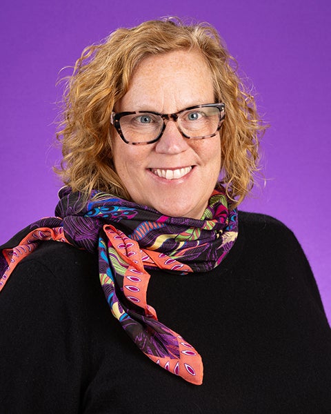 Dr. Megan Perry, with curly red hair, tortoise-shell glasses, and a colorful scarf and black sweater, smiles in front of a purple photo backdrop.