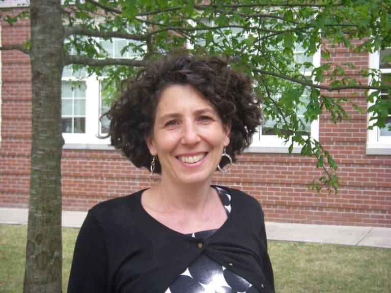 Dr. Laura Mazow, with curly dark hair, wearing a polka-dot blouse and black cardigan, stands smiling outside with a tree and a red brick building in the background.