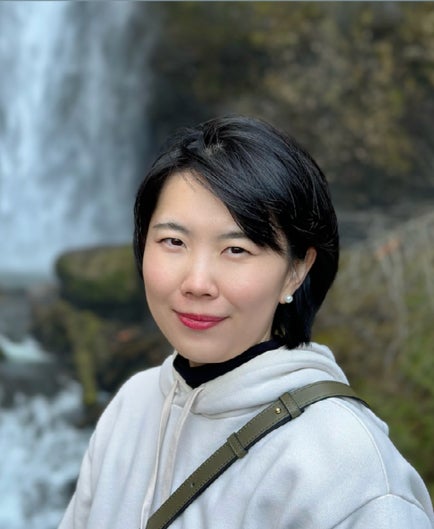 Dr. Yan Zhang, wearing a white pullover hoodie, has a green purse strap across her left shoulder and stands in front of a blurred background of green rocks and a waterfall.