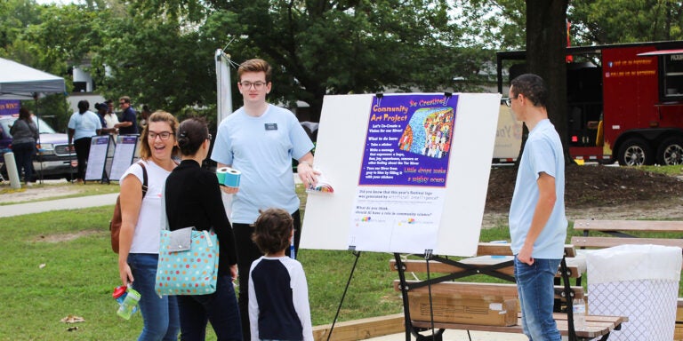 Local community families and children talk to an East Carolina University student at an exhibit outside at the Town Common in Greenville, North Carolina, during the 2023 Tar River Community Science Festival. A red food truck and tent with more staff appear in the background.