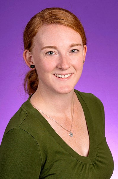 A woman with red hair pulled into a low ponytail is wearing a green v-neck shirt. She is photographed in front of a purple p backdrop.