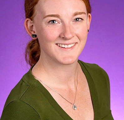 Dr. Erin Clark, with red hair pulled back in a low ponytail, is wearing an olive green v-neck shirt, and sits in front of a purple photo backdrop.