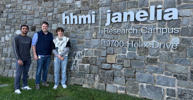 Three men in jeans and sweaters stand to the left of the HHMI Janelia Research Campus sign.