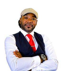 A man has his arms crossed and his head tilted. He is wearing a white shirt, black vest, red tie, and a beige cap.