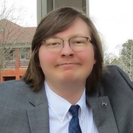 A man with shoulder-length brown hair is wearing glasses, a white shirt, a blue tie, and a gray suit jacket.