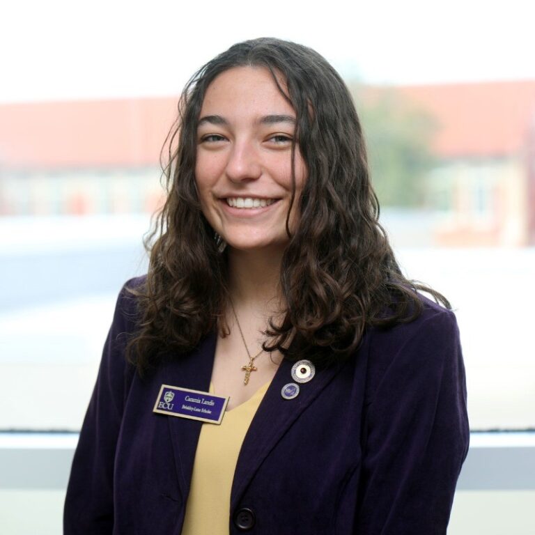 A woman with long, curly brown hair is wearing a black blazer over a tan shirt and has on an ECU nametag.