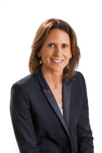 A woman with brown, shoulder-length hair is wearing a black suit jacket with a beige shirt underneath. She is sitting in front of a white background and smiling at the camera.