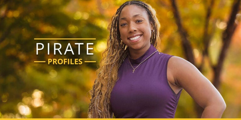 A woman in a purple tank top and long hair stands with her hand on her hip. The background contains trees with yellow, gold, and green fall-colored leaves.