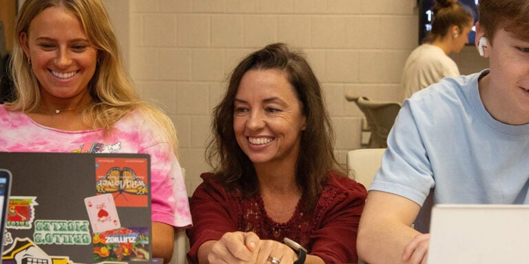 Two students on laptops sit on either side of a woman with long, brown hair who is kneeling between them with her hands on the table and looking at one of the laptop screens.