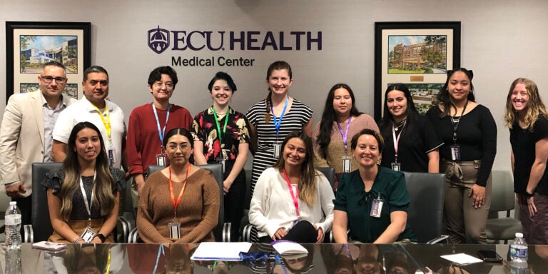 A group of 13 East Carolina University students, faculty and staff sit and stand at a long conference table with two photos and the words ECU Health Medical Center on the wall behind them.