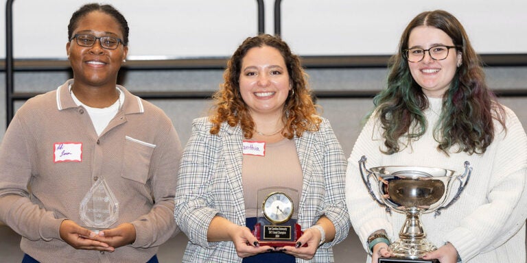 Three women stand in front of a white screen in East Carolina University’s Main Campus Student Center. They each hold a different trophy representing their first, second and third place awards in the Three-Minute Thesis competition.