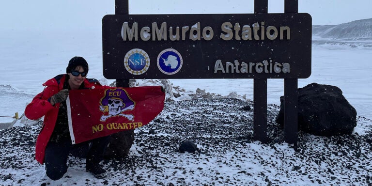 A man wearing winter outerwear holds a Pirate flag in front of the McMurdo Station sign in Antarctica.