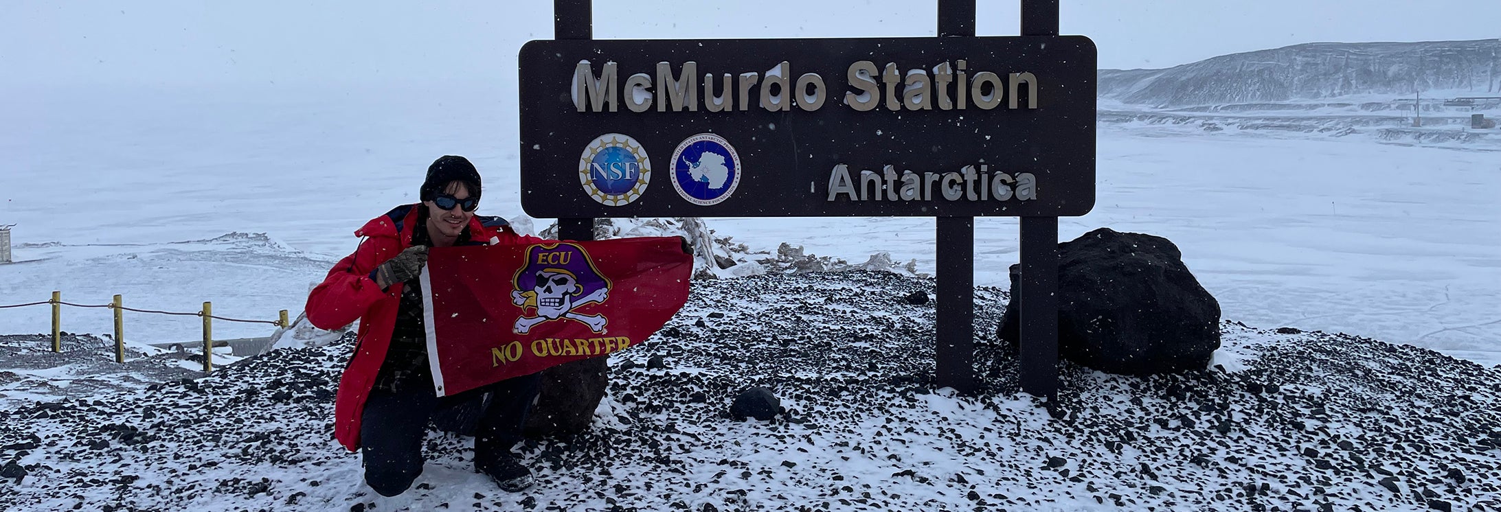 A man wearing winter outerwear holds a Pirate flag in front of the McMurdo Station sign in Antarctica.