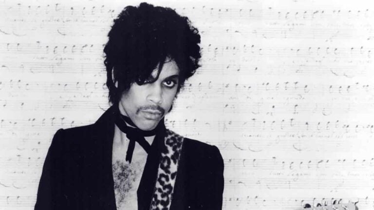 A black and white image of artist Prince standing in front of a background of sheet music.