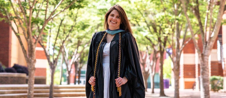 Biology staff member Ginger Grimes poses outside in front of trees lining a walkway on the East Carolina University campus while wearing a graduation gown and cords.