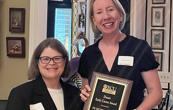 Two women in black shirts and name tags stand together with a plaque honoring the 2025 Harriot College Dean's Early Career Award recipient.