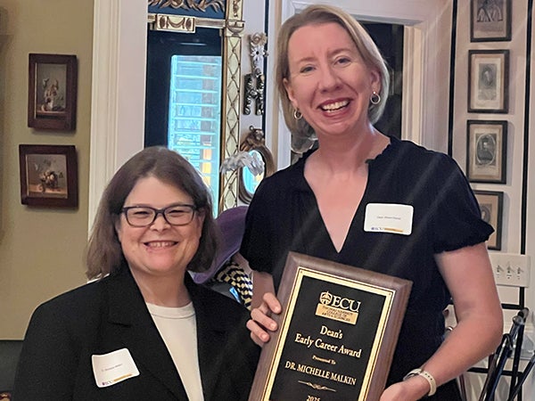 Two women in black shirts and name tags stand together with a plaque honoring the 2025 Harriot College Dean's Early Career Award recipient.