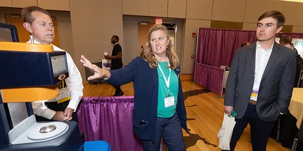 Two men in suits listen as a woman in a suit at an exhibit discusses a piece of equipment on a table.