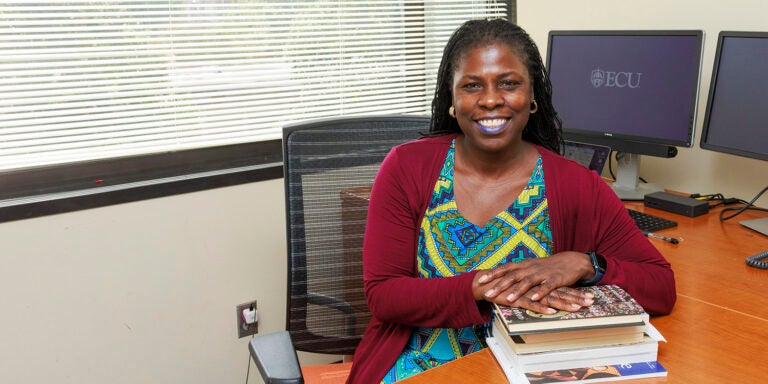 Dr. Marame Gueye sits at a desk with her arms on a stack of books and a computer monitor with the ECU logo in the background