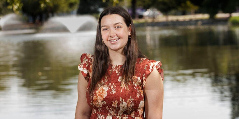 Female student with brown hair in a red, floral dress, with a pond in the background