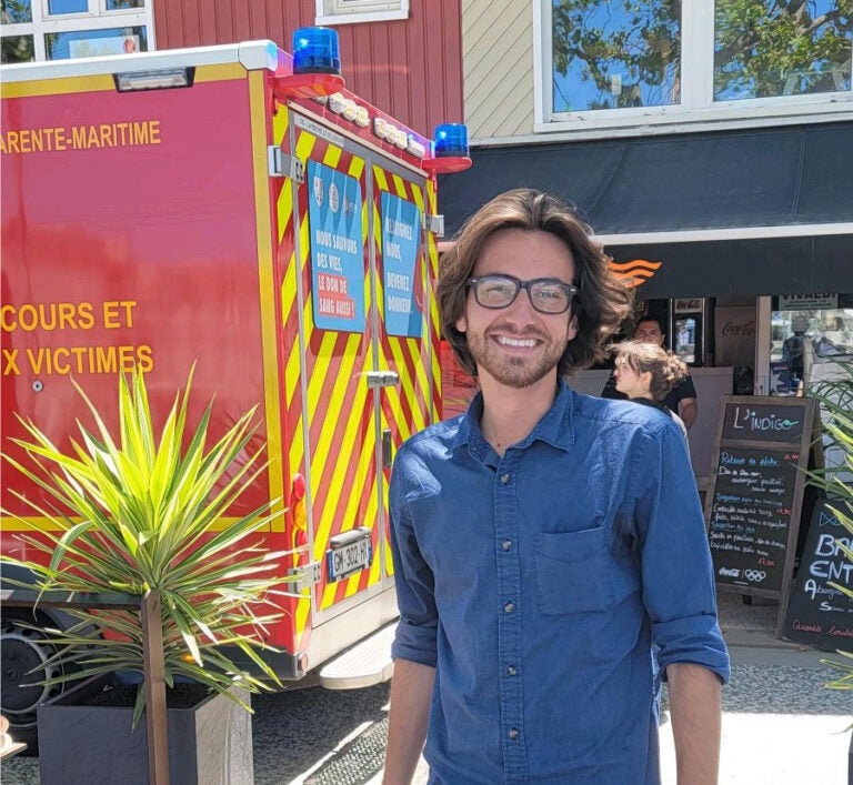 male student wearing glasses and a blue shirt stands smiling, in front of an ambulance.