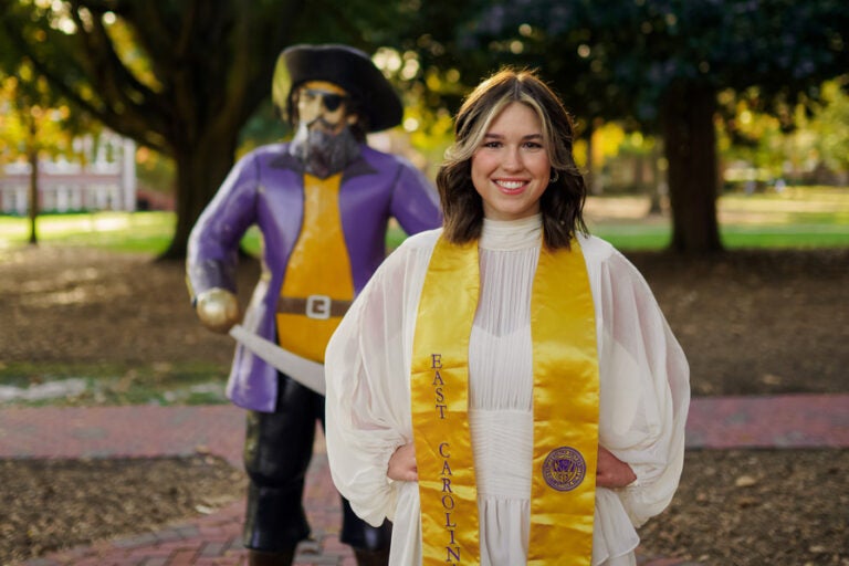 A women in a white dress with a gold sash stands in front of a Pirate statue on the East Carolina University campus.