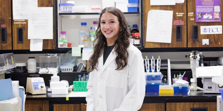 Bayli Locklear, wearing a white lab coat, stands in front of a countertop full of scientific equipment.