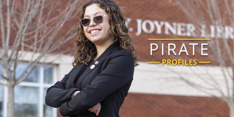 A female student with dark hair, sunglasses and a black blazer stands with arms crossed in front of Joyner Library.
