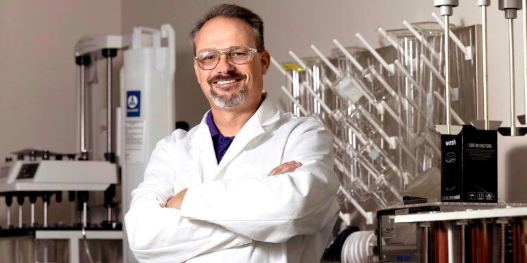 Man in a white lab coat stands in front of equipment in a lab