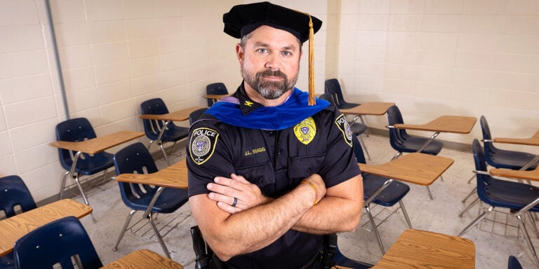 A man in a police uniform and a graduation cap with gold tassel stands with arms crossed in a classroom with desks.